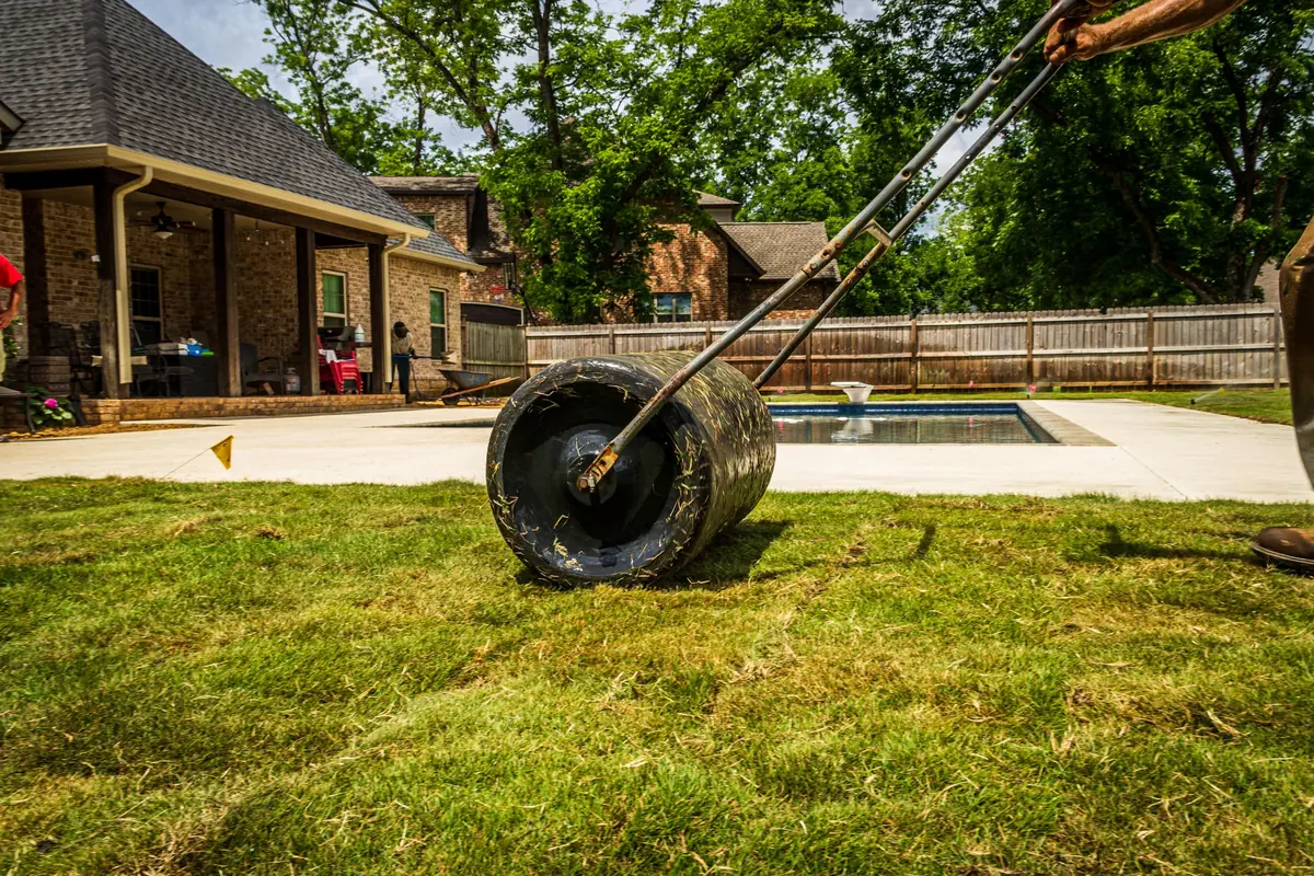 A lawn roller is pushed over newly laid instant lawn beside a paved pool surround.
