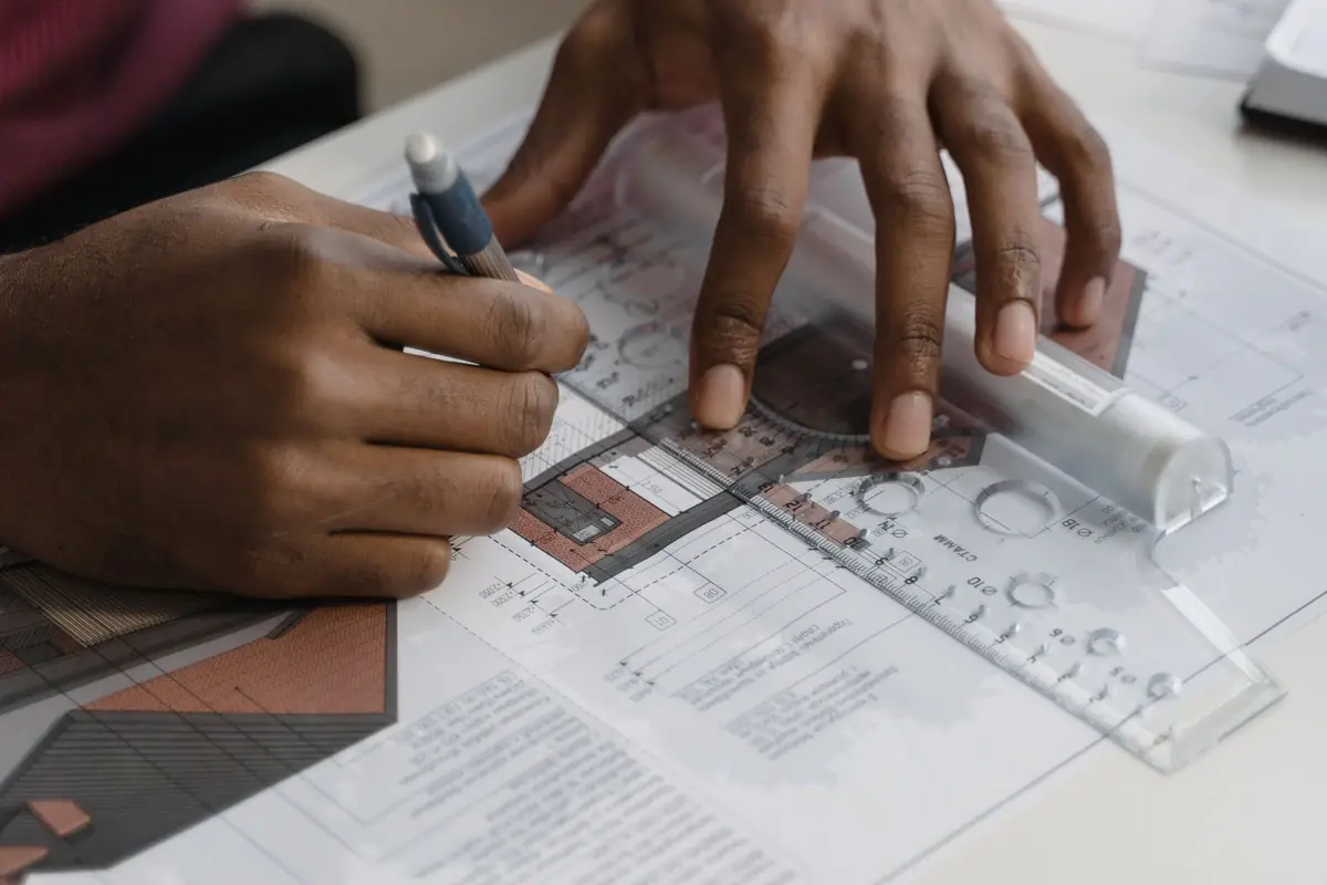 Hands mark up printed plans with a pencil and a drafting template.
