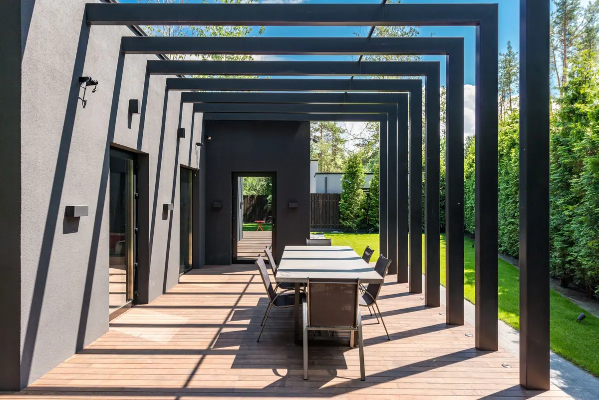 A timber deck with outdoor dining runs beside a lawn under black steel pergola frames.