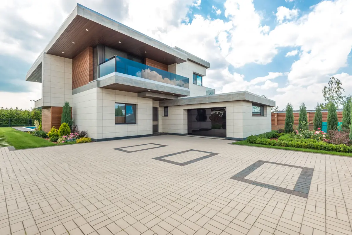 A light paved driveway with dark inlay squares leads between planted beds to a modern house.