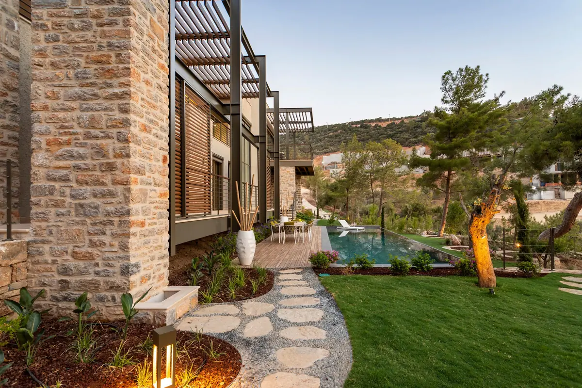 A stepping-stone path crosses a lawn between planted beds towards a pool and a stone-clad house.